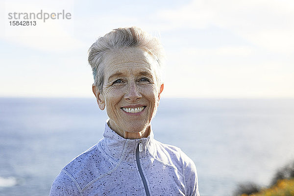 Porträt einer lächelnden älteren Frau  die bei Sonnenschein am Strand gegen den Himmel steht