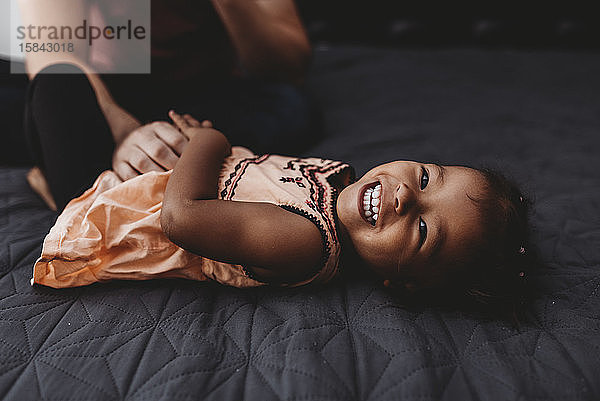 Süßes multirassisches 2 Jahre altes Mädchen liegt lachend auf dem Bett