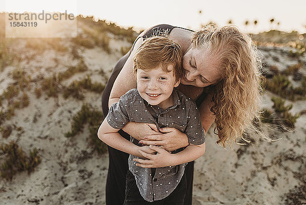 Frontansicht einer Mutter  die einen rothaarigen Sohn im Kindergartenalter bei Sonnenuntergang umarmt