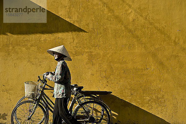 Vietnamesische Frau mit traditionellem vietnamesischen Hut trägt ihr Fahrrad
