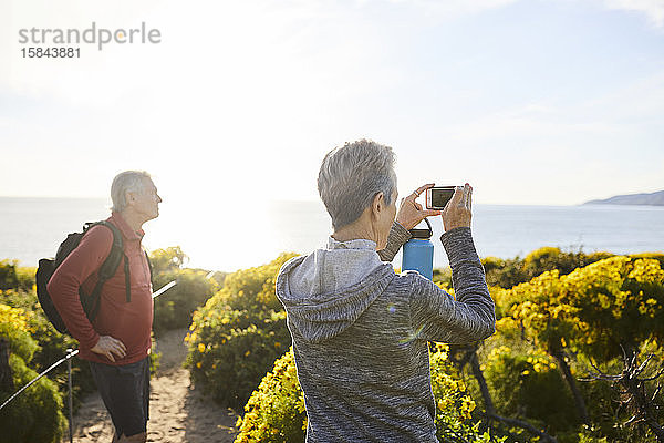 Rückansicht einer älteren Frau  die das Meer mit einem Smartphone fotografiert  während sie neben Pflanzen gegen den Himmel steht