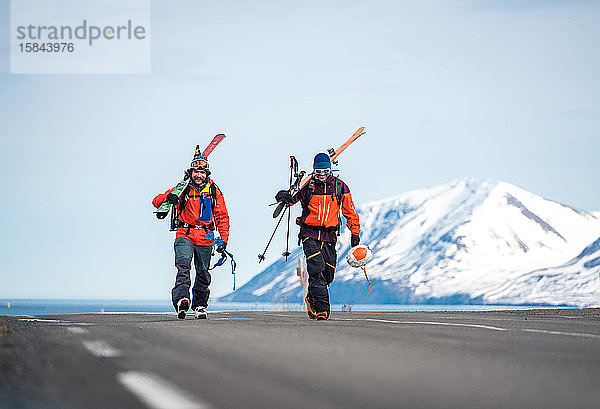 Zwei Männer gehen mit Skiern auf einer gepflasterten Straße in Island mit Bergen