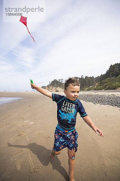 Junger Junge  der mit Entschlossenheit Drachen am Strand entlang läuft.