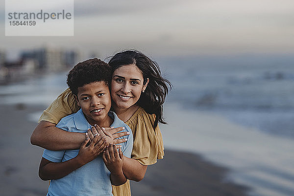 Nahaufnahme einer Mutter  die einen Jungen im Schulalter am Strand lächelnd umarmt