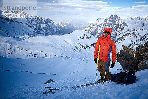 Skifahrer in orangefarbener Jacke  der sich auf Stöcke stützt und lacht