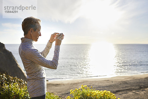 Ältere Frau fotografiert durch ein Smartphone  während sie an einem sonnigen Tag am Strand steht