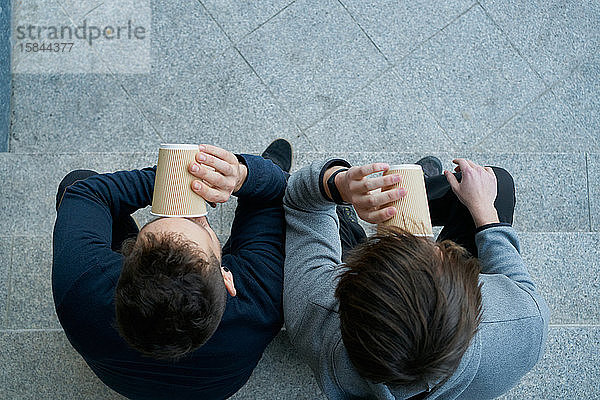Draufsicht auf zwei junge Männer  die auf einer Stadttreppe sitzen und Kaffee trinken