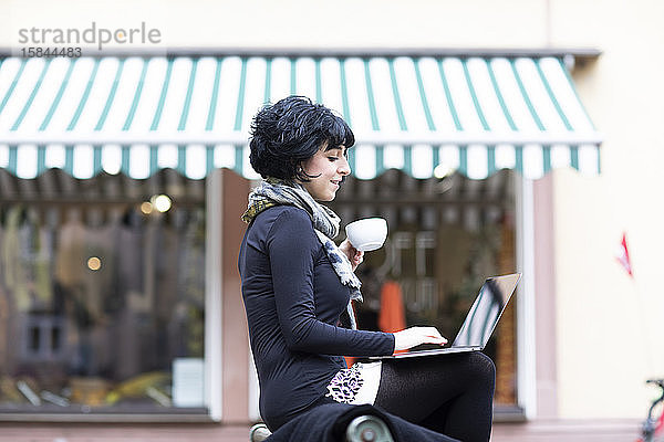 junge Frau  die in einem Laptop mit einer Kaffeetasse im Freien liest
