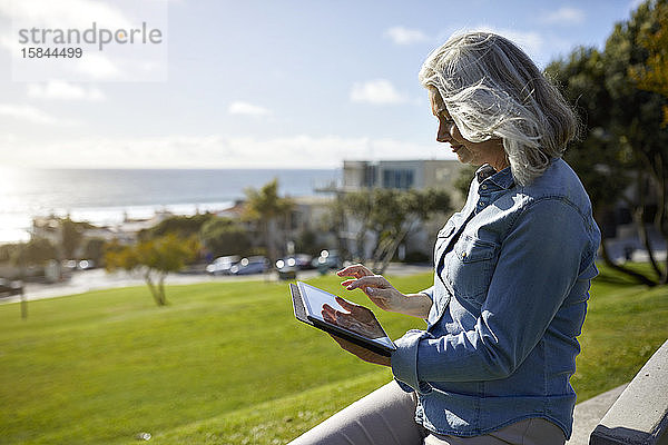 Seitenansicht einer Frau  die einen Tablet-Computer benutzt  während sie an einem sonnigen Tag auf einer Stützmauer sitzt