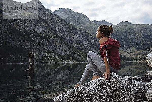 Frau mit Brötchen  die während einer Reise auf einem Felsen an einem See sitzt.