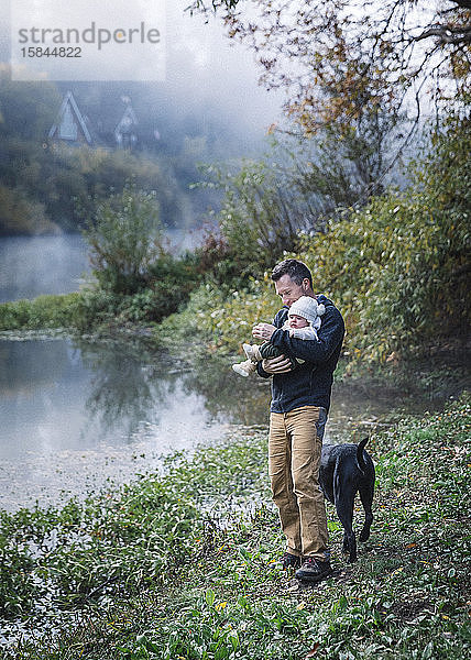 Ein Mann hält ein Baby in der Nähe eines Flusses und einen Hund