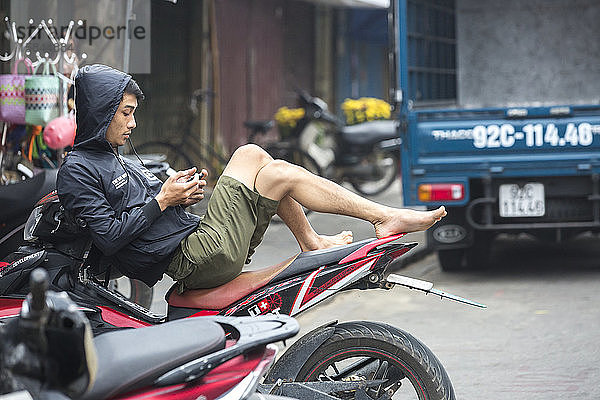 Vietnamesischer Mann entspannt sich auf Motorrad und schaut auf Smartphone.