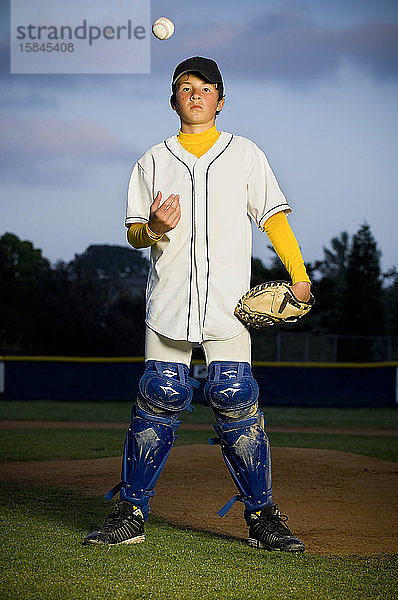 Porträt eines Jungen in weißer Baseball-Uniform und Schienbeinschützern  die einen Baseball in die Luft werfen