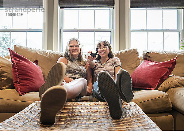 Zwei Mädchen im Teenageralter sitzen auf einer Couch mit den Füßen nach oben und schauen gemeinsam fern.