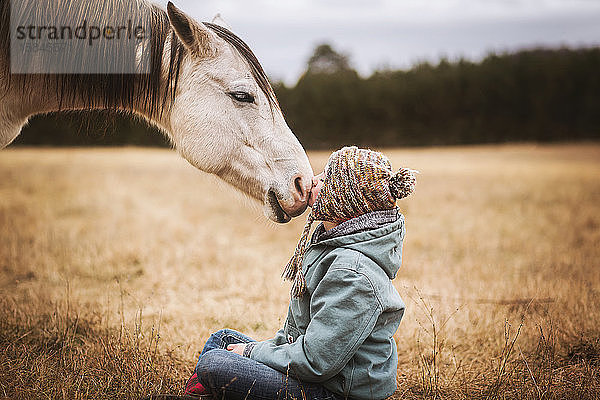 Junges Mädchen küsst weißes Pferd auf die Nase  während es am Boden sitzt