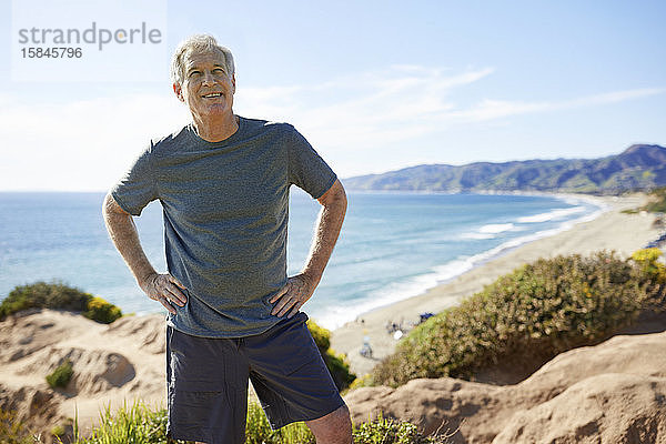 Lächelnder älterer Mann mit Händen auf den Hüften auf einer Klippe am Strand gegen den Himmel stehend