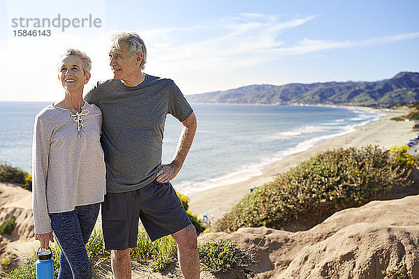 Lächelndes älteres Ehepaar steht am Strand auf einer Klippe gegen den Himmel während eines sonnigen Tages
