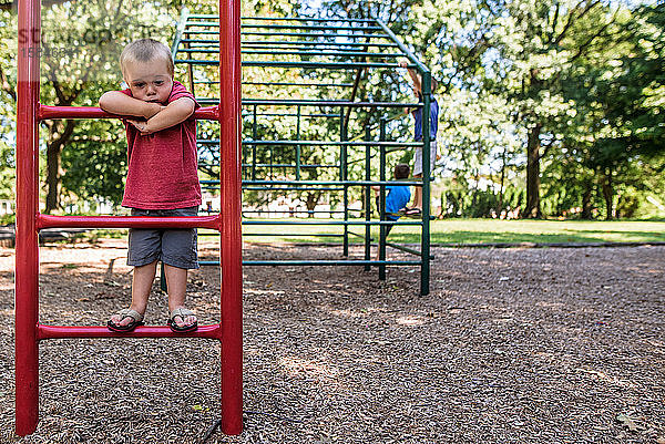 Junge sieht traurig auf dem Spielplatz aus  während ältere Jungen hinter ihm klettern