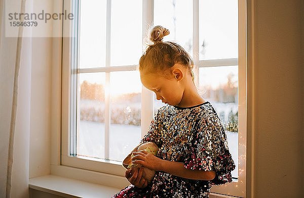 junges Mädchen  das zu Hause bei Sonnenuntergang ihr Spielzeug in ein glitzerndes Kleid kuschelt