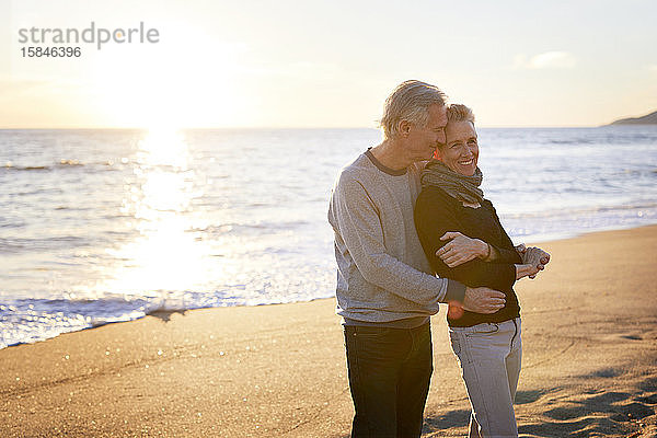 Zärtliches älteres Ehepaar  das bei Sonnenuntergang am Strand gegen den Himmel steht