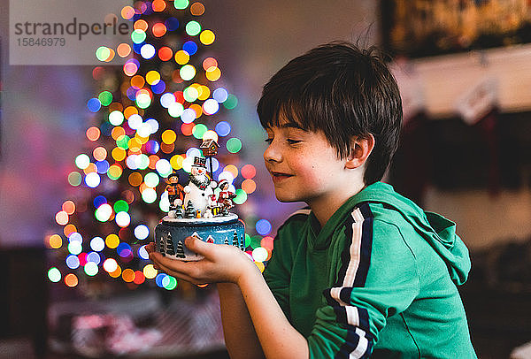 Junge hält Weihnachtsschmuck mit Baumlichtern im Hintergrund.