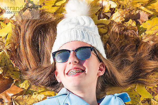 Teenager-Mädchen mit Sonnenbrille und Mütze liegt auf dem Boden  bedeckt mit gelben Blättern  während sie den sonnigen Tag genießt. Das brünette Mädchen ist glücklich und lächelt. Horizontales Foto