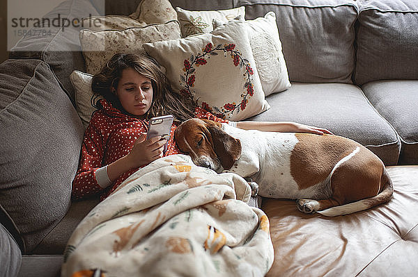 Tweener-Mädchen schaut auf ihr Telefon  während sie mit dem Jagdhund auf der Couch kuschelt