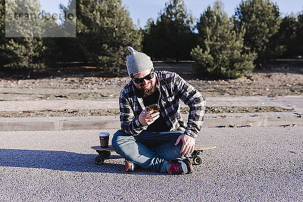 Moderner junger Mann sitzt auf einem Longboard und schaut auf sein Mobiltelefon
