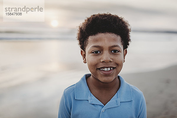 Porträt eines jungen Jungen im Schulalter  der bei Sonnenuntergang am Strand lächelt