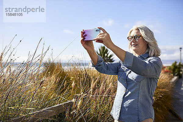 Ältere Frau fotografiert mit Smartphone  während sie am Strand von Manhattan gegen den Himmel steht