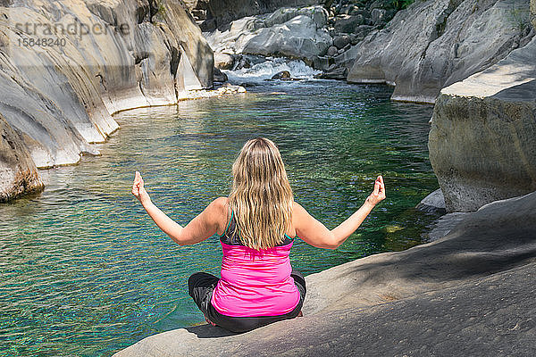 Frau rückwärts in sportlicher Kleidung  die an einem idyllischen Ort neben einem Fluss und einem Wasserfall zwischen weißen Felsen entspannende Yoga-Übungen praktiziert. Der Yogi sitzt mit erhobenen Armen auf dem Boden