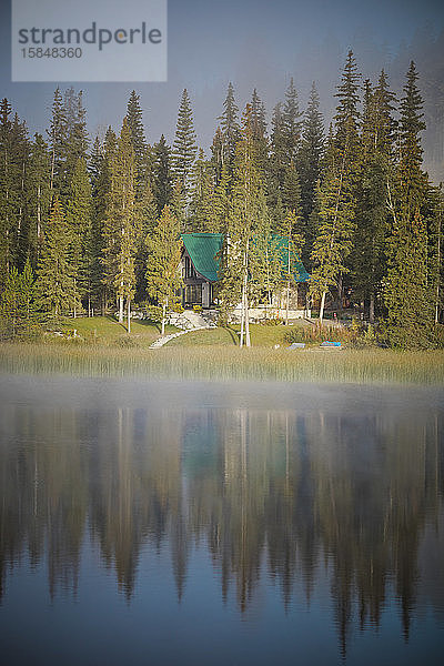 Hütte mit grünem Dach  das sich im ruhigen See spiegelt.