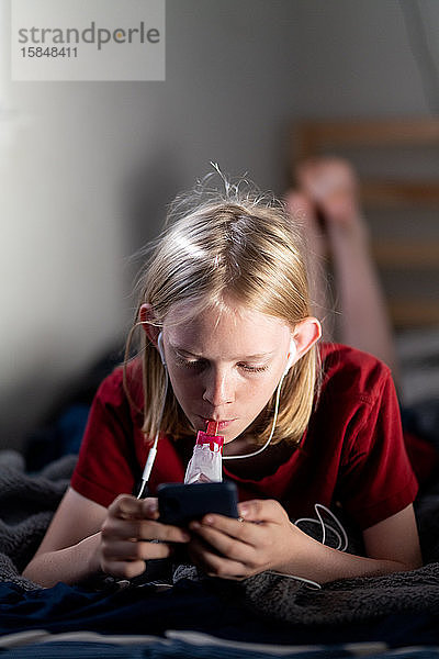 Junge Person  die auf dem Bett liegt und mit Ohrstöpseln mit Eis am Stiel auf das Telefon schaut