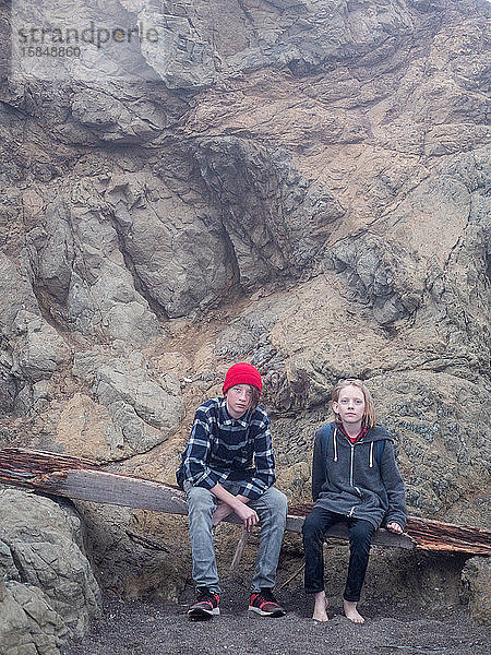 Teenager-Brüder sitzen auf Treibholz am Fuße einer Felsklippe