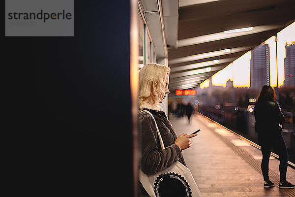 Junge Frau wartet am U-Bahnhof mit einem Smartphone auf den Zug