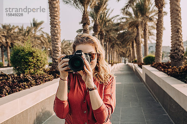 Junge Frau  die in einer Palmenallee steht und mit ihrer Kamera fotografiert