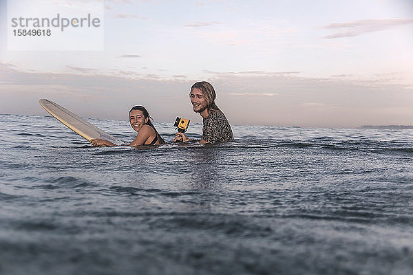 Glückliches Paar mit Surfbrett und Kamera im Meer