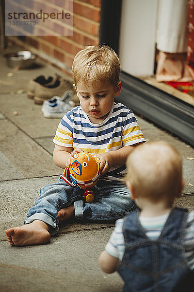 Kleinkind sitzt draußen und spielt mit Spielzeug  beobachtet vom kleinen Bruder