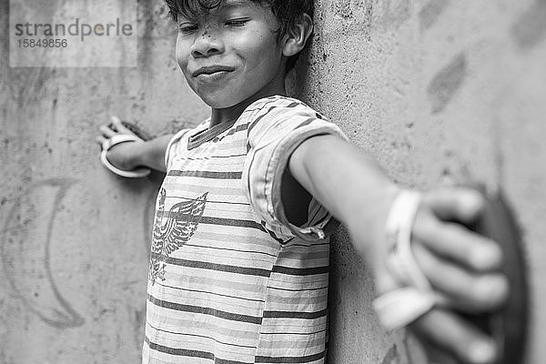 Lächelnder brasilianischer Indianer  der mit Pantoffeln in den Händen an einer Wand lehnt