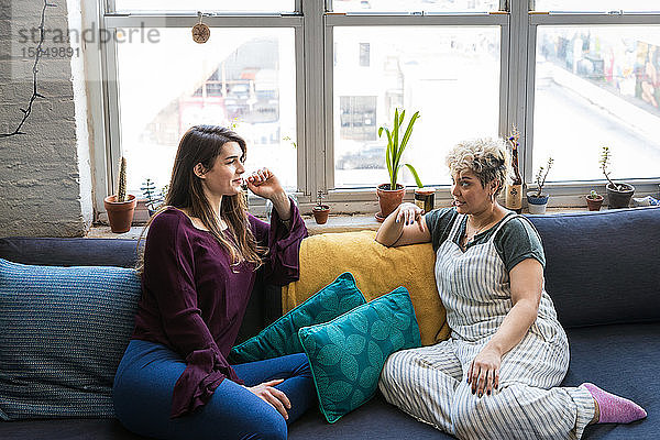 Weibliche Mitbewohnerinnen unterhalten sich  während sie sich zu Hause auf dem Sofa am Fenster entspannen