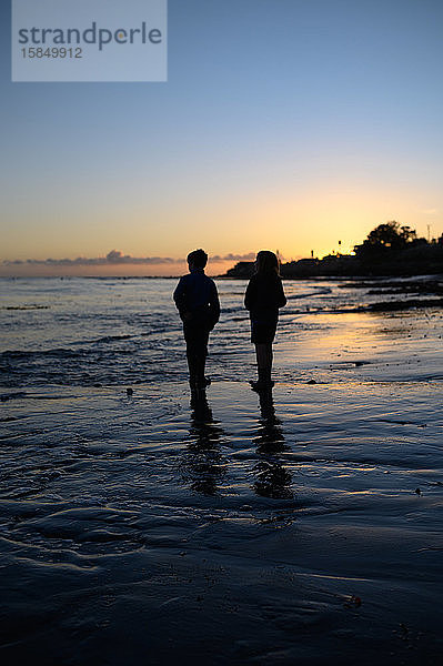 Silhouette von zwei Kindern  die bei Sonnenuntergang am Strand stehen