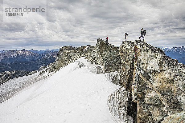 Drei Bergsteiger stehen auf einem felsigen Gipfel über einem schneebedeckten Gletscher.