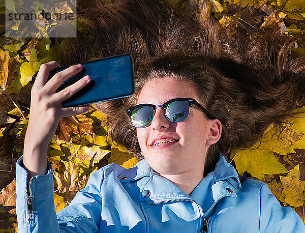 Teenager-Mädchen  das eine Sonnenbrille trägt und mit gelben Blättern bedeckt auf dem Boden liegt  während sie ein Foto mit ihrem Mobiltelefon für sich selbst macht. Das Mädchen lächelt in die Kamera. Horizontales Foto