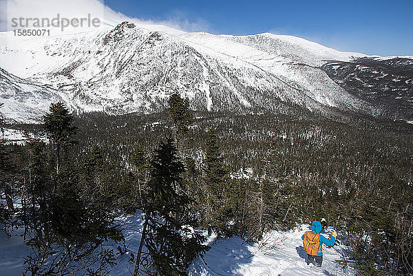 Eine Frau  die allein einen verschneiten Hang hinunterwandert  mit der Tuckerman-Schlucht und dem Gipfel des Mount Washington im Hintergrund.