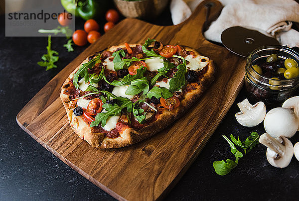 Nahaufnahme einer handgemachten Pizza auf Holzbrettern mit Zutaten drum herum.