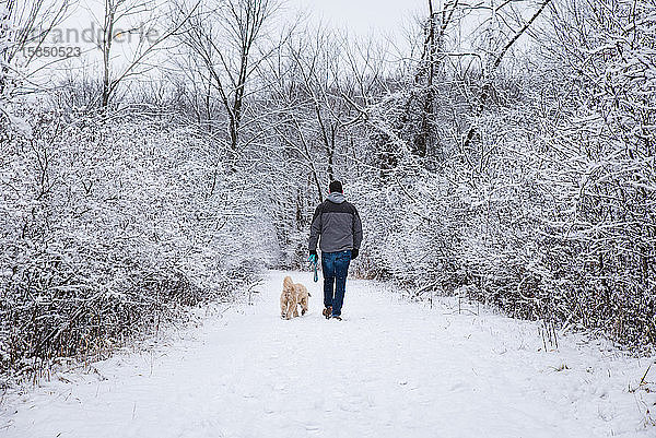Mann  der an einem Wintertag mit einem Hund auf einem verschneiten Pfad durch den Wald spaziert.