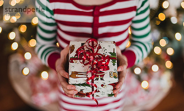 Kind im Pyjama hält Weihnachtsgeschenk vor Lichter am Baum.