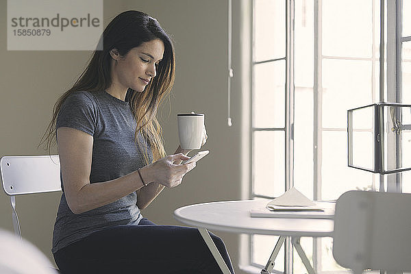 Frau benutzt Mobiltelefon  während sie im Wohnzimmer Kaffee am Tisch am Fenster trinkt