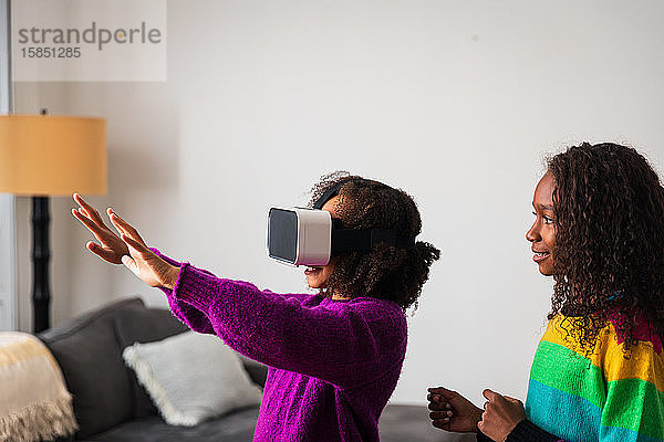 Lächelndes Mädchen sieht Schwester beim Gestikulieren an  während sie zu Hause einen Virtual-Reality-Simulator trägt