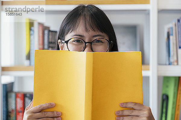 Fröhliche junge Frau deckt Gesicht mit gelbem Textbuch.